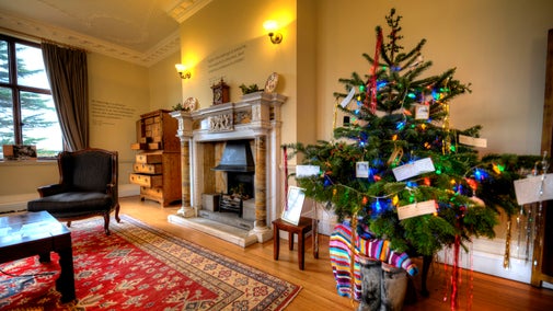 A christmas tree on the right of the image decorated with coloured lights. A pair of wellies and a bright striped jumper are at the bottom of the tree. There is a fire place to the left and then a leather arm chair to the left of the fire place